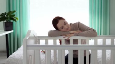 Yorgun genç anne yeni doğan bebeğin yattığı karyolaya yaslanarak uykuya daldı. Annelik kavramı. Annem doğum izninde. Yeni doğmuş bir bebeğe bakma kavramı. Yavaş çekim.