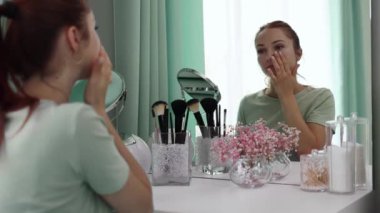 Genç bir kadın aynaya yavaş çekimde bakarken yüzüne krem sürüyor. Cilt bakımı. Güzellik anlayışı, kişisel bakım ve günlük krem kullanımı.