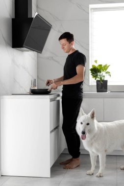 Genç bekar bir adam mutfakta köpeğiyle yemek pişiriyor. Beyaz İsviçreli Çoban. Evcil hayvanlarla yaşama kavramı. Ev rutini.