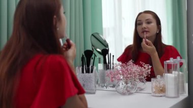 Gece elbisesi giymiş bir kız dudaklarına ruj sürer. Makyaj yapan genç ve çekici bir kadın tuvalet masasının arkasında ağır çekimde.