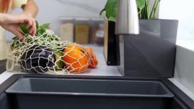 Genç bir kadın, modern mutfakta organik sebze ve meyveleri tekrar kullanılabilir bir poşetten yavaş çekimde çıkarır. Market alışverişi. Sıfır israf. Eko-Arkadaş Alışverişi. Sağlıklı yaşam tarzı kavramı.