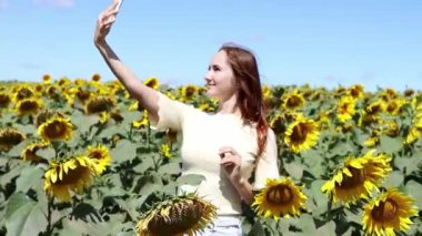 Genç bir kız akıllı telefon kullanıyor, cep telefonu ekranına bakıyor, selfie çekiyor ve ayçiçeği tarlasında yavaş çekimde bir video görüşmesi yapıyor. Çevrimiçi iletişim kavramı. Bağımsız.
