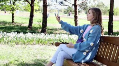 Genç bir kız parkta bir bankta oturuyor ve akıllı telefon kullanıyor, cep telefonuna bakıyor, selfie çekiyor ve cep telefonuyla yavaş çekimde bir video görüşmesi yapıyor. Çevrimiçi iletişim kavramı. Bağımsız.
