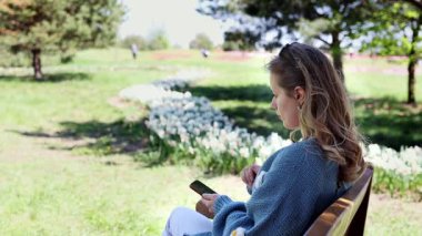 Genç bir kız parkta bir bankta oturuyor ve akıllı telefon kullanıyor, cep telefonuna bakıyor, selfie çekiyor ve cep telefonuyla yavaş çekimde bir video görüşmesi yapıyor. Çevrimiçi iletişim kavramı. Bağımsız.