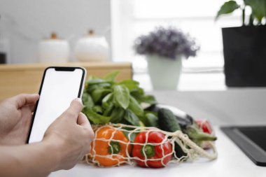 Yeniden kullanılabilir bir çantada taze sebze ve meyvelerin yanında beyaz ekran bir telefon tutuyor. Yemek servisi. Model-up. İnternetten market alışverişi. Sıfır israf. Sağlıklı bir yaşam tarzı fikri