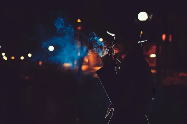 man detective agent in a hat and raincoat smokes a cigarette at night in a rainy city in the style of film noir