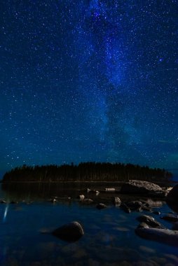 Yıldızlı bir gökyüzü ve parlak yıldızlı Samanyolu ile gece manzarası. Astrofotografi takımyıldızları ve galaksiler ile bir gölün suları üzerinde kayalar ve arka planda bir orman.
