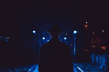 Kara film tarzında yağmurlu bir şehirde gece vakti şapkalı ve yağmurluk giyen bir dedektif ajan.