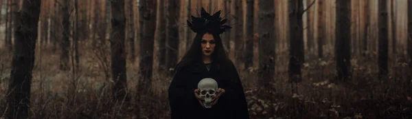 森で死んだ男の手に頭蓋骨を持った黒い恐ろしい魔女 バナーヘッドカバーのためのパノラマの広い横の写真 — ストック写真