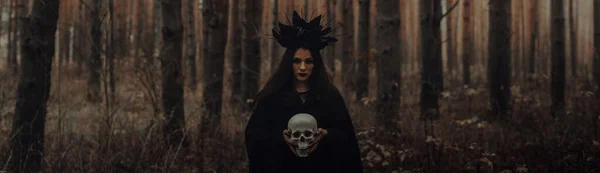 一个死在森林里的人手里拿着骷髅的可怕的黑巫婆 横幅头盖全景横向照片 免版税图库图片