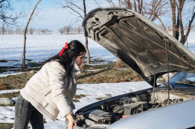 Kız, kışın yolda bozuk bir arabanın motoruna bakar.