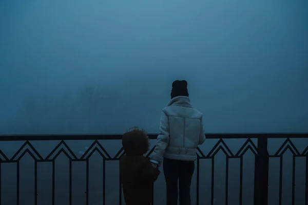 Backs of a woman with a child boy on a misty foggy bridge on an autumn evening in the fog mist