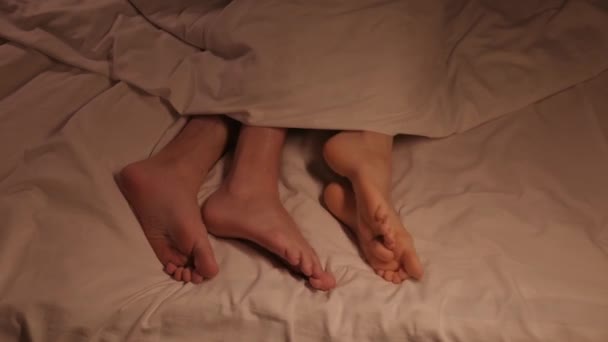 夜の自宅の寝室のベッドの上の白いシートの毛布の下でセックスをしているカップルの足 男と女が愛を作っている — ストック動画