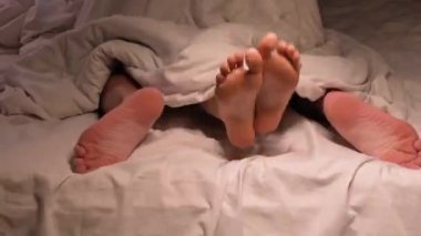 Bir çiftin bacakları battaniyenin altında yatak odasındaki beyaz çarşafın üzerinde sert seks yapıyor. Bir erkek ve bir kadın sevişiyor.