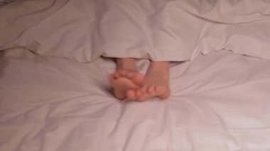 Çocuk bacakları, geceleri battaniyenin altında parmaklarıyla dans eder ve eğlenir. Çıplak ayaklı mutlu kız bebek geceleri yatak odasındaki rahat yatakta uyumuyor.