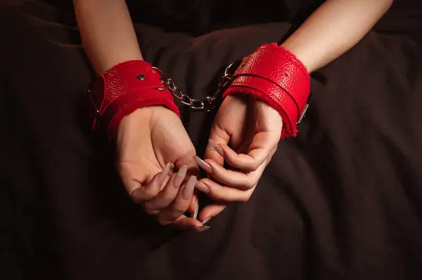 female hands in red leather BDSM handcuffs on a black sheet in close up. Submissive woman slave girl is lying on bed in the bedroom. Sex with submission and domination