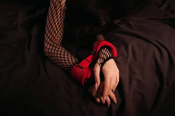 female hands in red leather BDSM handcuffs on a black sheet in close up. Submissive woman slave girl is lying on bed in the bedroom. Sex with submission and domination