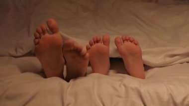 Oğlan ve anne ayaklarının dans eden ayakları çarşafın altında. Mutlu kız bebek kız ve kadın anne yatak odasında çıplak ayakla oynaşıyor.