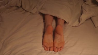 Kadın bacakları neşeyle film çeker ve yatakta battaniyenin altında yatarken ayak parmaklarıyla dans eder. Çıplak ayaklı mutlu kadın dinleniyor ve akşamları bir otel odasında dinleniyor.