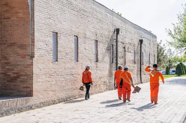 stock image group of Uzbek male cleaners janitors in orange uniforms with shovels outside Barak Khan Madrasah in spring. Tashkent, Uzbekistan - April 17, 2024