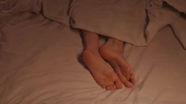 Kadın bacakları neşeyle film çeker ve yatakta battaniyenin altında yatarken ayak parmaklarıyla dans eder. Çıplak ayaklı mutlu kadın dinleniyor ve akşamları bir otel odasında dinleniyor.