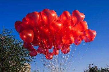 Mavi gökyüzünün arka planında kalp şeklinde bir sürü kırmızı balon var.