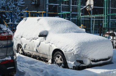 Otoparka karla kaplı bir araba park edildi. Arabanın lastikleri, tekerlekleri ve kaputu beyaz bir kaplamayla kaplı.