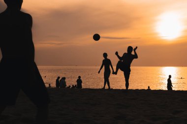 Gün batımında deniz kenarında geleneksel plaj futbolu oynayan bir grup Asyalı adamın siluetleri.