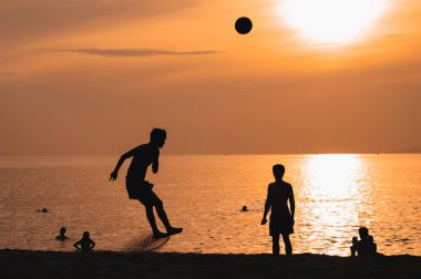 Sahilde güneş doğarken plajda futbol oynayan Asyalı erkeklerin siluetleri.
