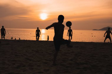 Gün batımında deniz kenarında geleneksel plaj futbolu oynayan bir grup Asyalı adamın siluetleri.