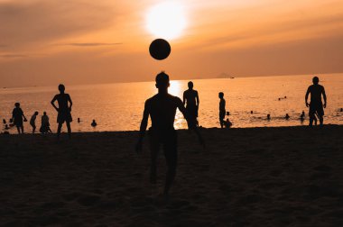 Gün batımında deniz kenarında geleneksel plaj futbolu oynayan bir grup Asyalı adamın siluetleri.