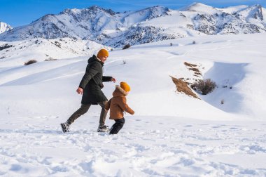 Baba ve oğul kışın bir dağ manzarasının arka planında karla kaplı bir yamaçta koşuyorlar..