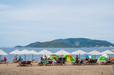 Yazın bulutlu bir günde plaj şemsiyeleri altında Vininci Adası 'na bakan kumlu bir sahilde güneşli yataklarda tatil yapan turistler. Nha Trang, Vietnam - 21 Temmuz 2024