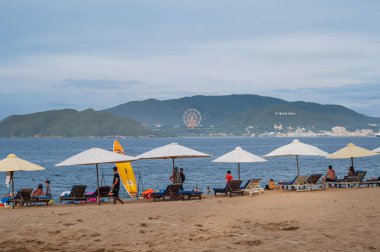 Yaz akşamları Vininci Adası 'na bakan kumlu bir sahilde kumsal şemsiyeleri altında güneşli yataklarda tatil yapan turistler. Nha Trang, Vietnam - 21 Temmuz 2024