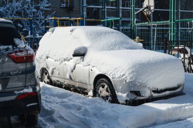 Otoparka karla kaplı bir araba park edildi. Arabanın lastikleri, tekerlekleri ve kaputu beyaz bir kaplamayla kaplı.