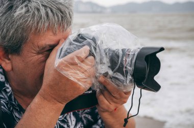 Profesyonel erkek fotoğrafçı, dijital kamerayla koruyucu bir çantada ve su geçirmez plastik torbayla doğada yağmur damlalarına karşı fotoğraf çeker.