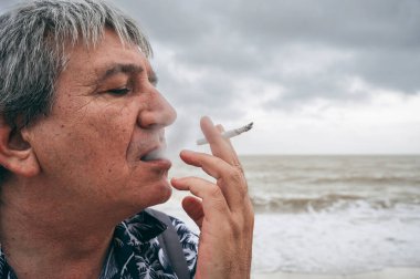 Yalnız, üzgün, yaşlı, yaşlı beyaz bir adam sahilde sigara içiyor.