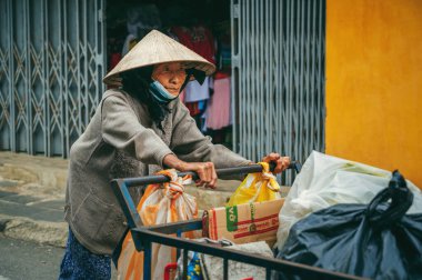 Yaşlı üzgün Asyalı Vietnamlı kadın çöp toplayıcı Hoi An, Vietnam 'da sokakta çöp arabası taşıyor. Hoi An, Vietnam - 2 Aralık 2024