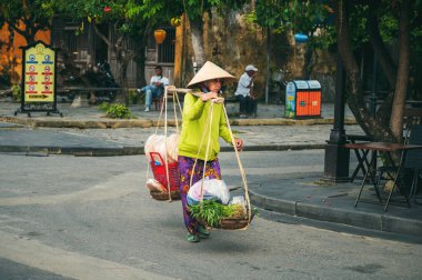 Geleneksel Non La şapkalı Vietnamlı kadın satış elemanı Asya 'da Vietnam' da Hoi An şehrinin sokaklarında sepet içinde yemek satıyor. Hoi An, Vietnam - 2 Aralık 2024