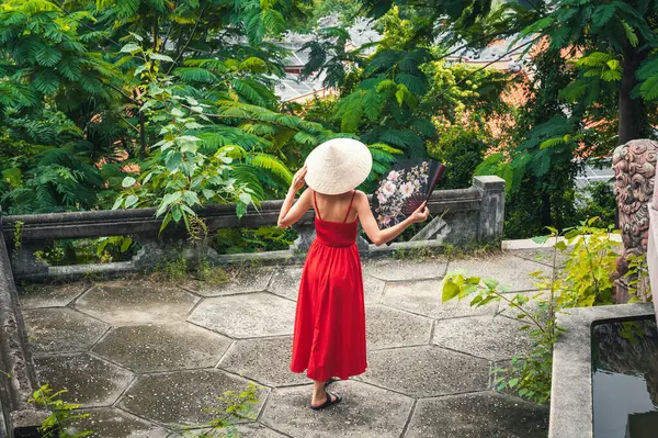 Vietnam 'da bir bahçede yelpazesi olan Vietnamlı non-la şapkalı güzel bir turist kadının arkasında. Asya 'da turizm ve seyahat