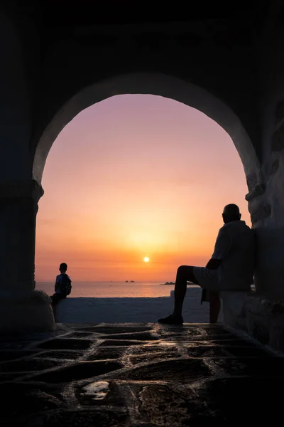 Unknown boy and old man looking at the sea at sunset from a Greek arch