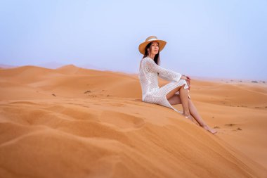 Deve şapkalı ve beyaz elbiseli genç esmer kadın Sunset, Fas 'ta Erg Chebbi Dunes' da oturuyor.