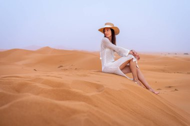 Deve şapkalı ve beyaz elbiseli genç esmer kadın Sunset, Fas 'ta Erg Chebbi Dunes' da oturuyor.