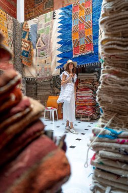 Marakeş, Fas 'taki bir goblen dükkanında beyaz elbiseli ve şapkalı genç siyah saçlı kadın poz veriyor.
