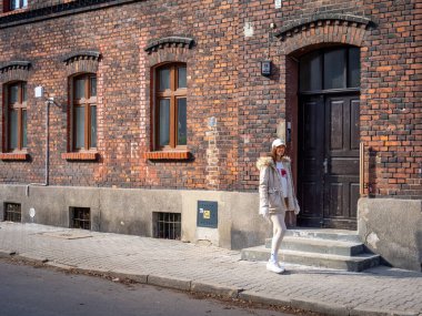 Polonya 'nın güneşli bir kış gününde Katowice' deki kırmızı tuğlalı binanın önünde bej ceket ve şapka giyen genç bir manken. Yüksek kalite fotoğraf