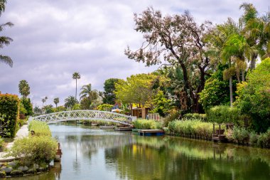 Los Angeles, Kaliforniya 'da Venedik' te bulutlu gökyüzüne karşı kurulmuş büyüleyici bir köprü ve renkli bitkiler içeren huzurlu bir kanal manzarası
