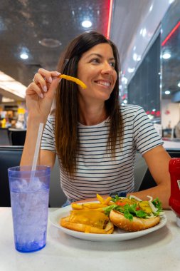 Bir kadın lokanta masasında oturmuş lezzetli bir hamburgerin tadını çıkarırken yanında kızarmış patates kızartması yiyor.