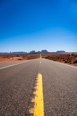 Geniş çölde dolambaçlı uzun bir yol, merkezinde belirgin sarı bir çizgi ile işaretlenmiş, ABD 'deki Anıt Vadisi' nin ana yolundaki kurak arazide gezginlere rehberlik ediyor.