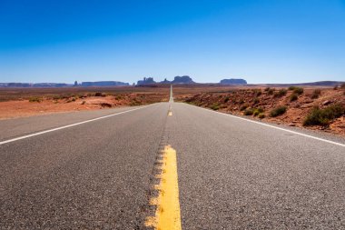 Engin çöl boyunca uzanan uzun, dolambaçlı bir yol, arka planda belirgin bir şekilde yükselen görkemli dağlarla, ABD 'deki Anıt Vadisi' nin ana yolunda.