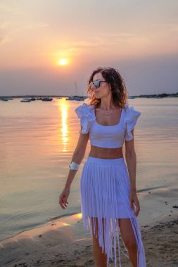 Beyaz elbiseli şık bir kadın gün batımında plajın kenarında durur, Formentera, Balearic Adaları 'ndaki suda parlayan yansımalarını yakalar..